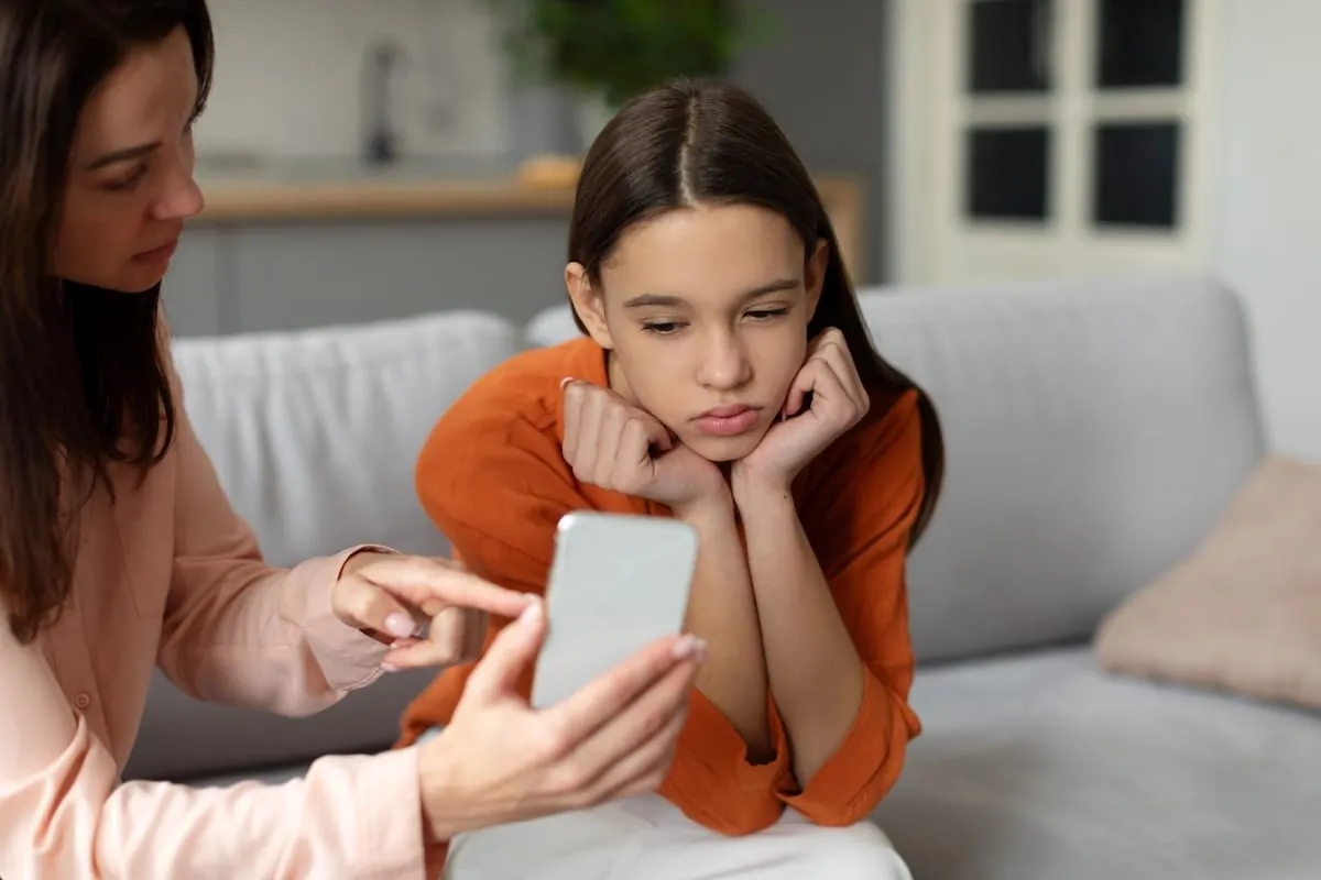 Mother holding a smartphone in front of her teenage daughter, who sits on the couch with her chin in her hands looking unhappy